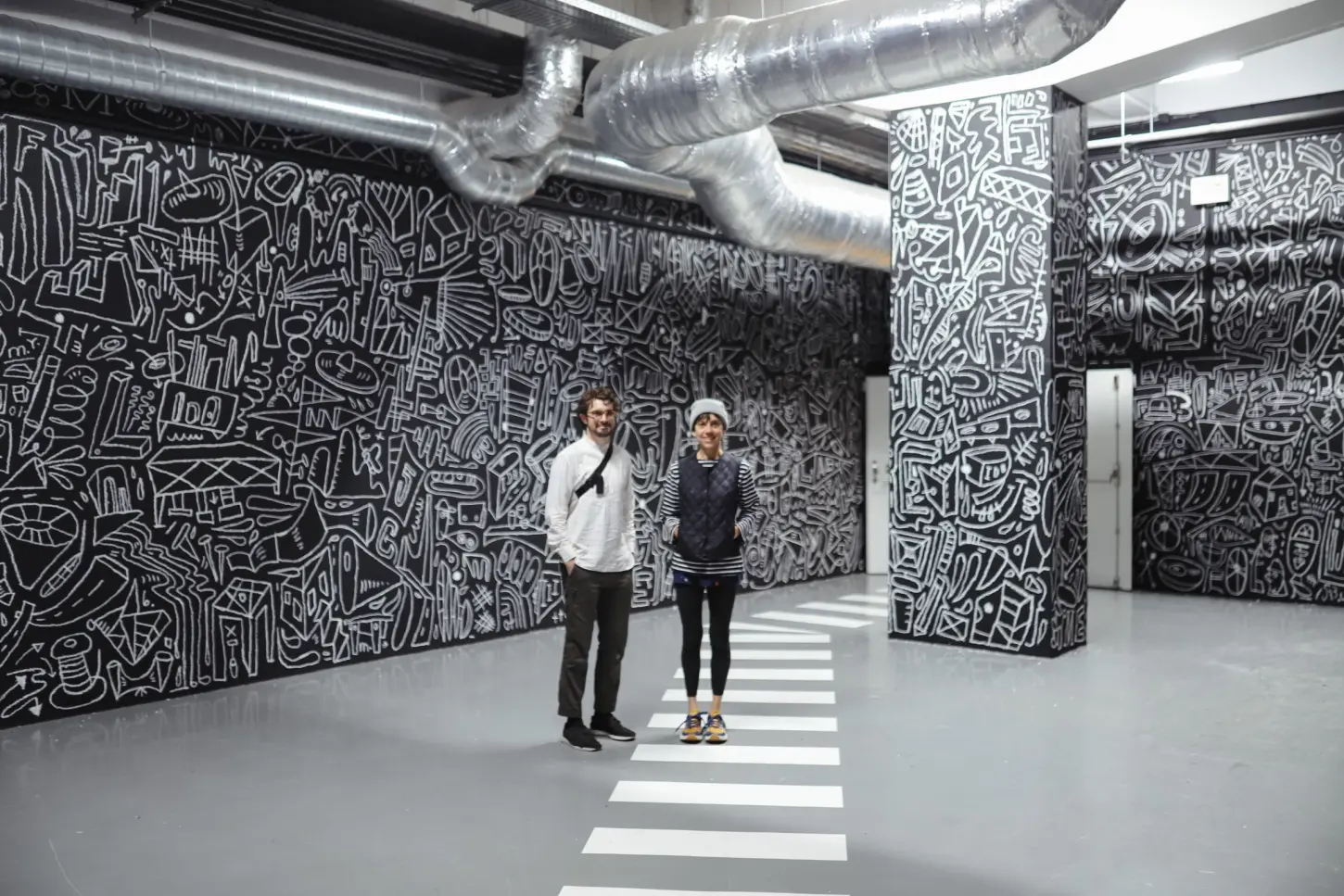 a man and woman standing in a room with a wall with black and white drawings